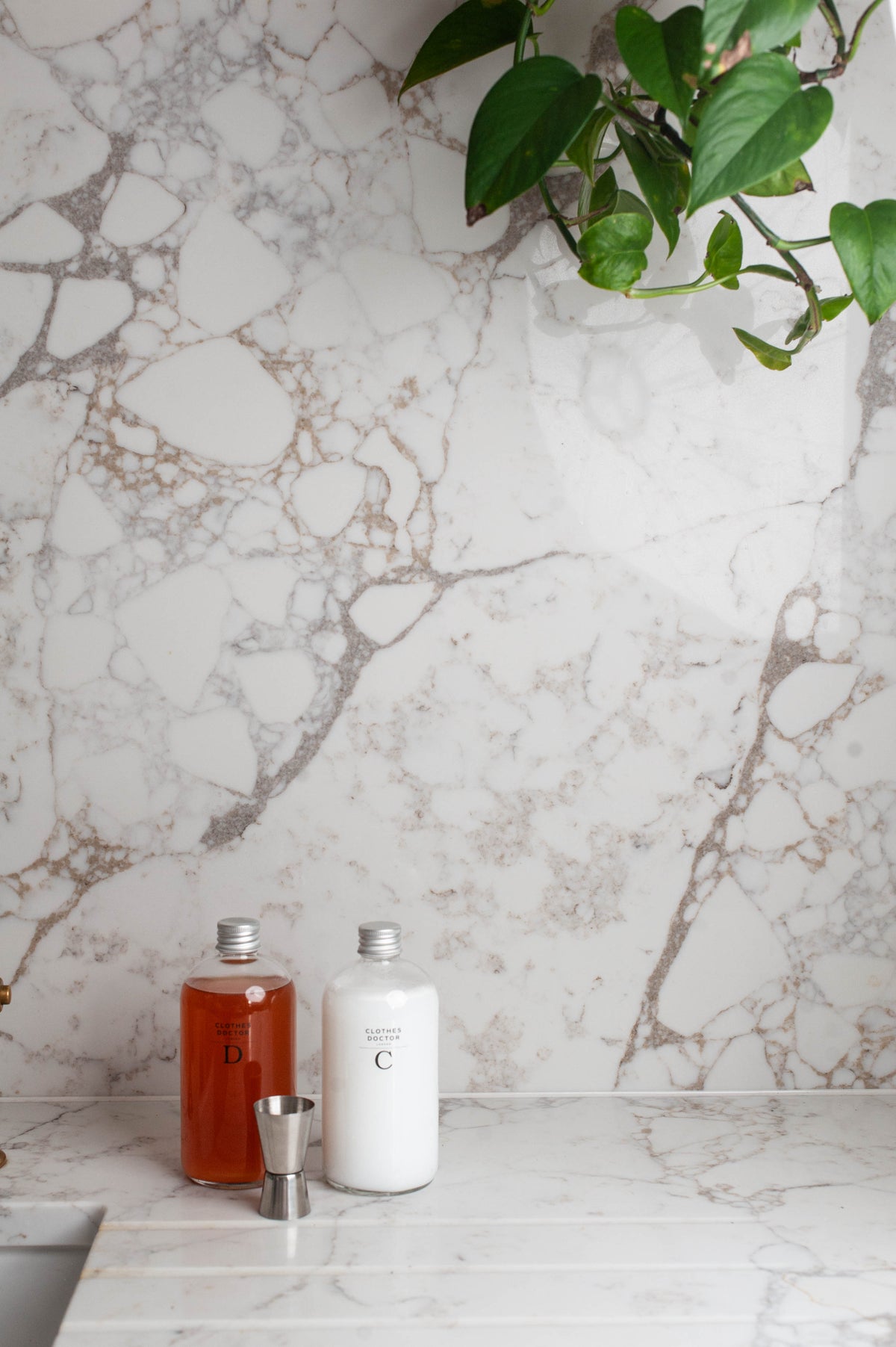 Two minimalist bottles, one amber liquid and one white liquid, with a small metal jigger, set on a white marble countertop against a matching marble backsplash with a green plant hanging in the top right.
