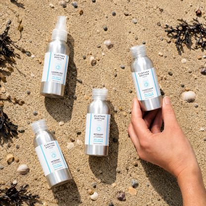 Four silver spray bottles of Clothe Doctor seawater treatment arranged on sandy beach with shells and seaweed, with a hand holding one bottle.