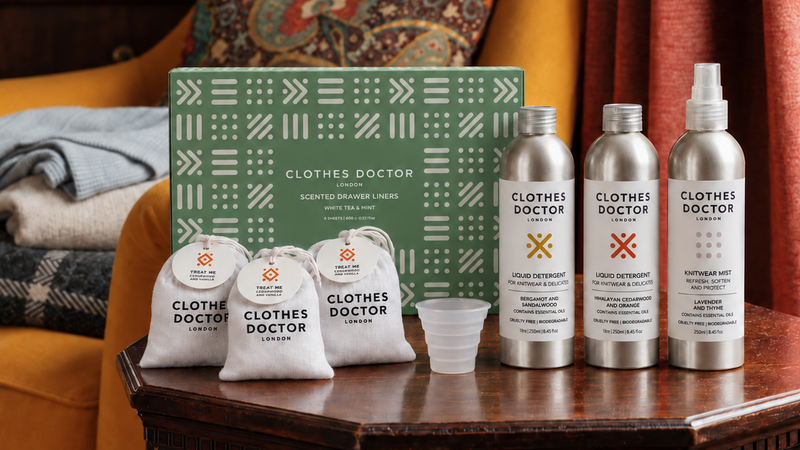 Clothes Doctor scent restoration bundle displayed on a dark wooden table in a cozy living room setting. The bundle includes a green patterned box, three silver bottles of liquid restoration, three white drawstring pouches, and a small clear plastic cup.